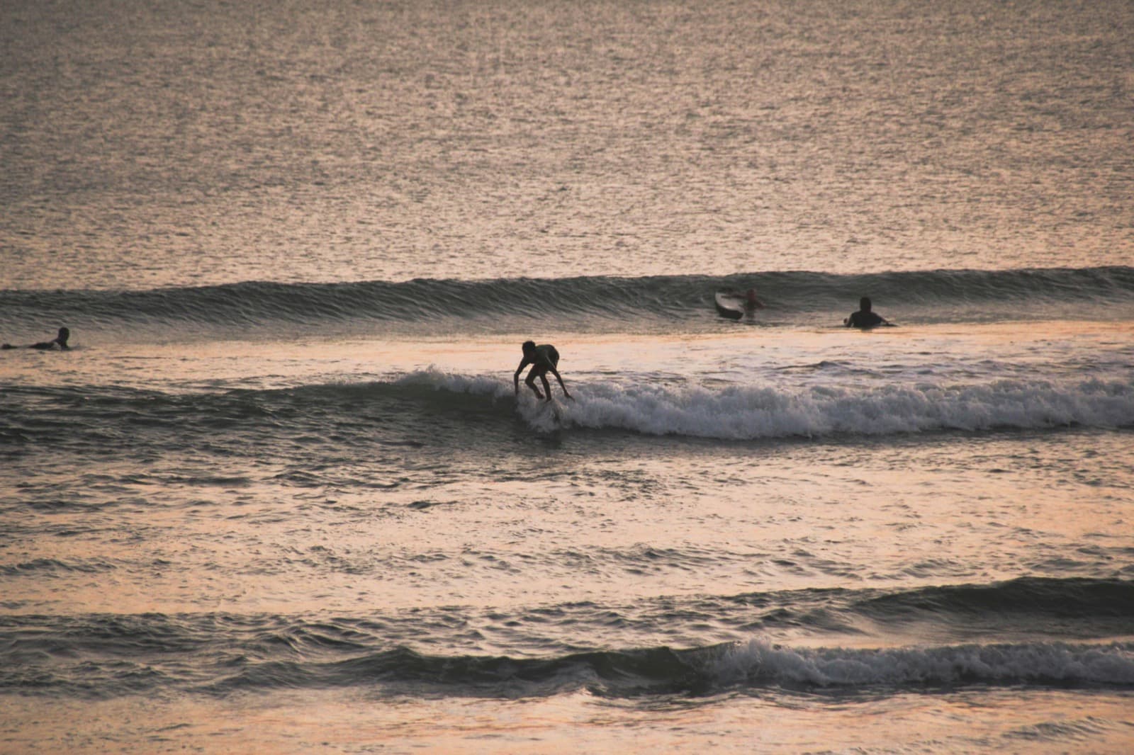 Seminyak Beach at sunset, golden hour on Bali's south coast, couples and surfers silhouetted against the fading light