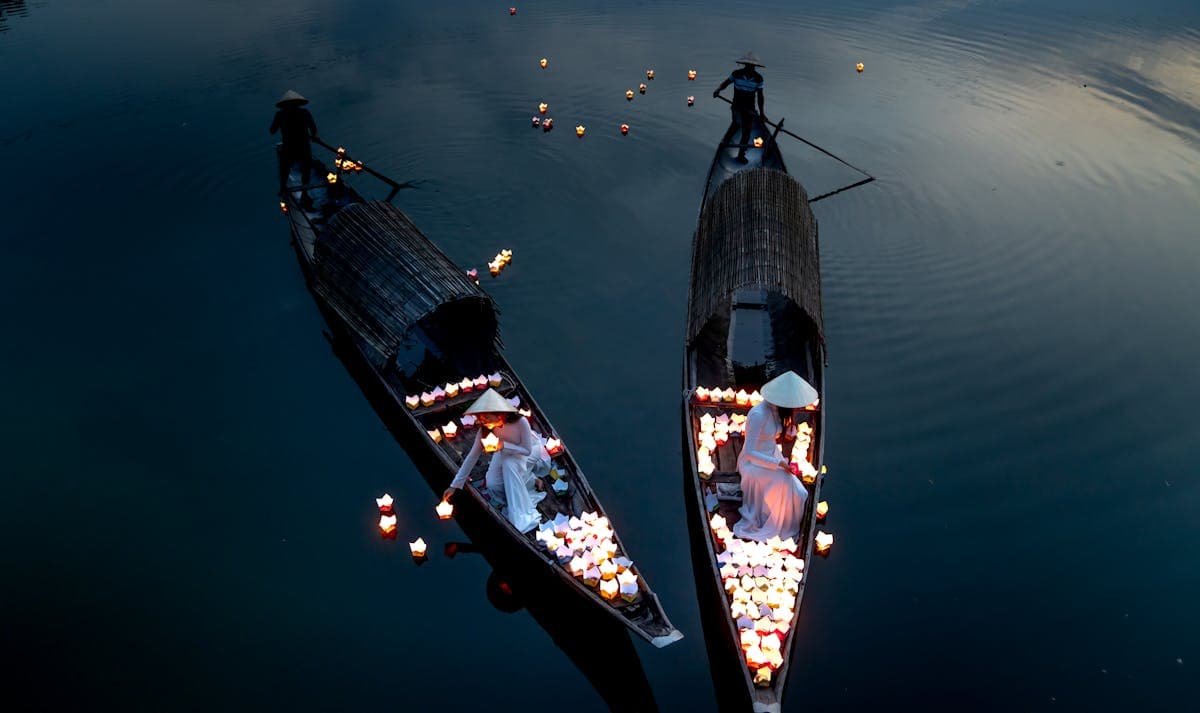 Lantern boats on the Thu Bon River at dusk, women in white ao dai float paper lanterns on the water. This aerial scene is Hoi An at its most magical on full-moon nights.