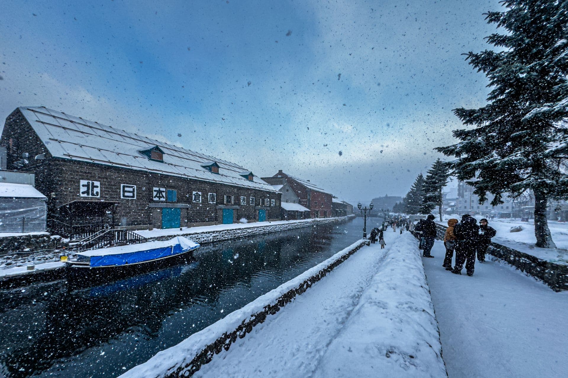 Daytime snowfall over the Otaru Canal and its stone warehouses, with visitors on the promenade