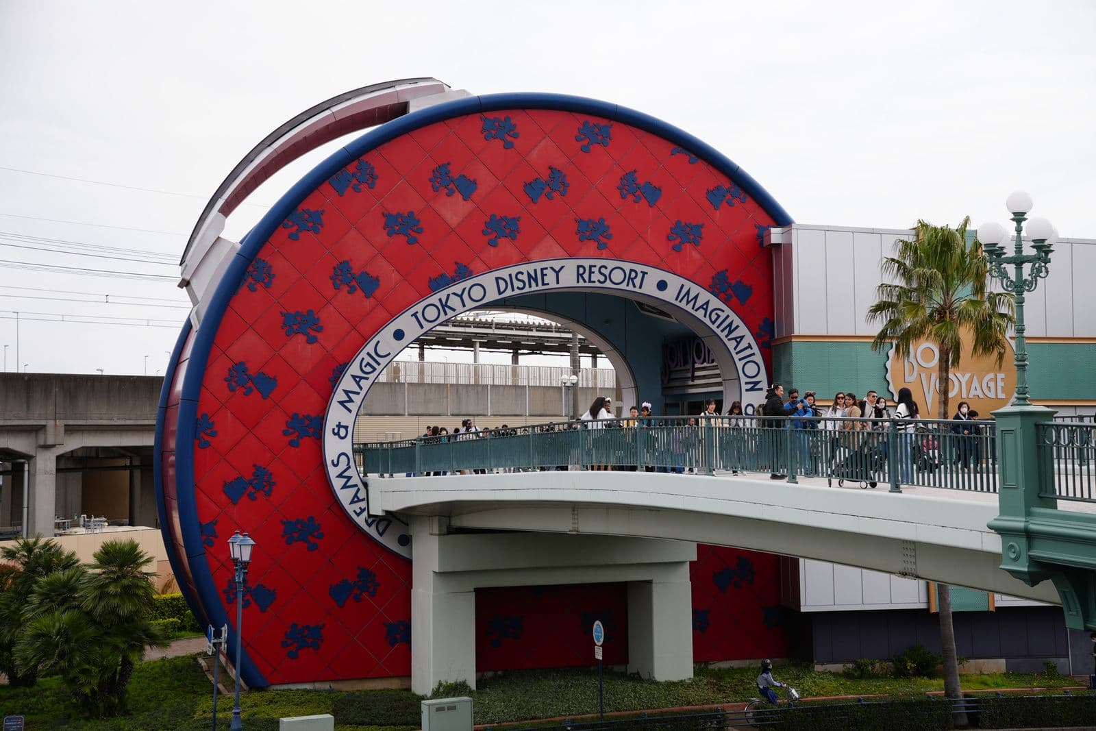 TOKYO DISNEY RESORT 문구가 둘린 빨간 원형 게이트와 보행교, 오른쪽에 봉 보야지 매장이 보이는 리조트 입구